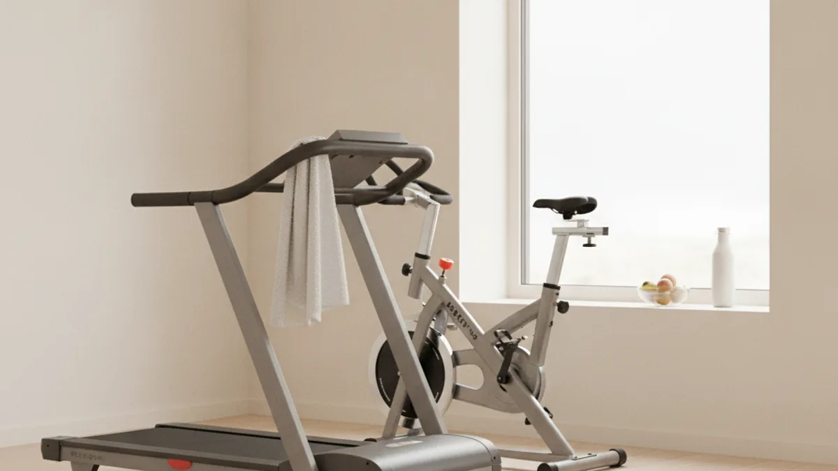 A treadmill with a towel on the handrail and an exercise bike by a large window in an empty room, with a bottle and a bowl of fruit on the windowsill