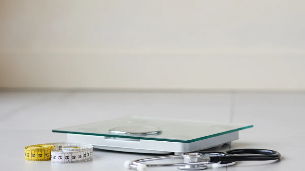 Glass floor scales on a light-colored floor, next to a coiled yellow-and-white measuring tape and a stethoscope with black tubes