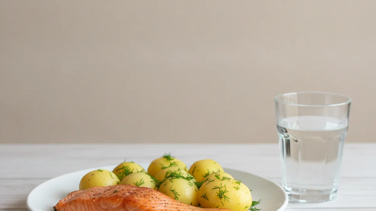 Тарелка с запечённой рыбой, отварной картофель и стакан воды на светлом кухонном столе