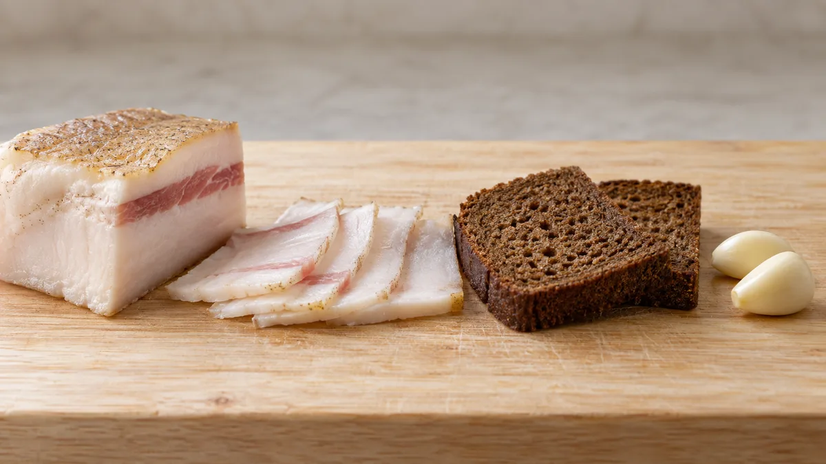 A block of salted salo with a streak of meat, thin slices of salo, rye bread and garlic on a wooden board